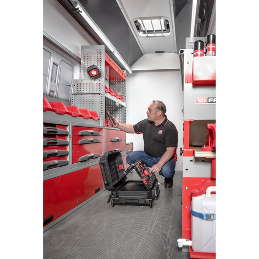 A person kneeling inside a well-organized mobile workshop van with red and grey Facom cabinets and drawers. The person is handling tools from a black rolling case and appears to be organizing or checking the storage units.
