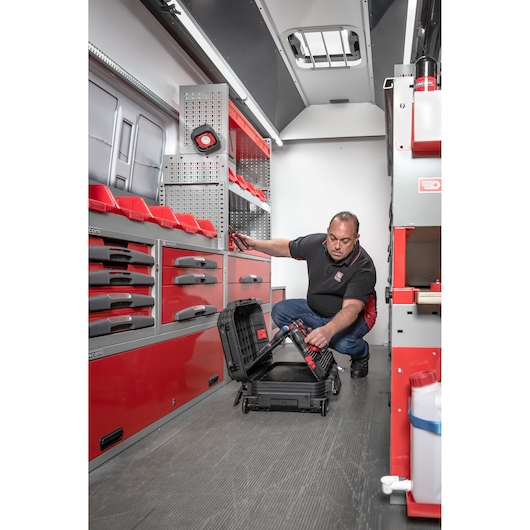 A person is kneeling inside a workshop van, organizing tools into a black hard case. The van is equipped with red and grey drawers, bins and tool storage compartments. The person’s face is intentionally blurred for privacy.