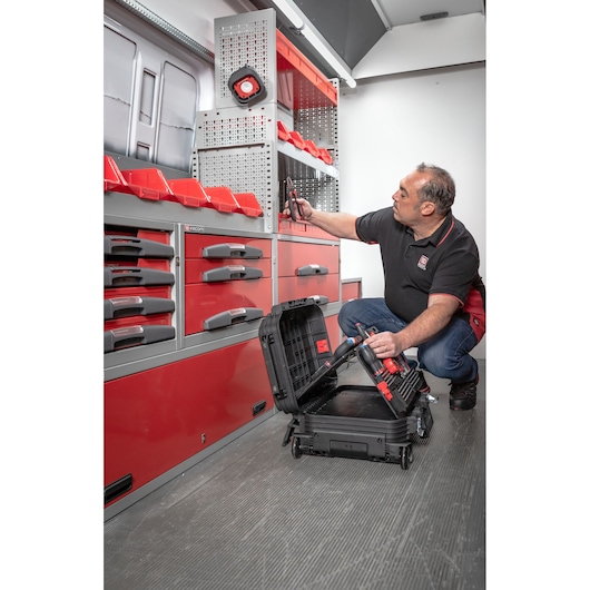 A person is kneeling inside a workshop vehicle, organizing tools using a portable Facom tool case. The vehicle interior features red and grey storage units, shelves with bins, and pegboards for tool organization.