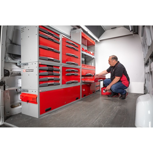 A man is kneeling inside a van fitted with red and grey Facom tool storage units. He is opening a drawer and holding a small organizer case, surrounded by organized tool drawers and shelves.