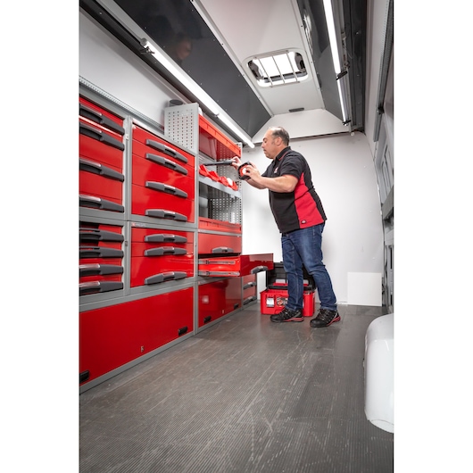 A person standing inside a vehicle workshop with red Facom tool cabinets and drawers, organizing tools. The workspace is well-lit and features various storage units for tools.