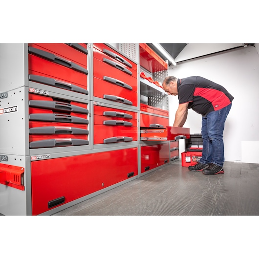 A person wearing a black and red shirt is opening a red drawer in a storage unit with multiple red and grey drawers labeled Facom. The scene takes place in a workspace with organized tool storage.