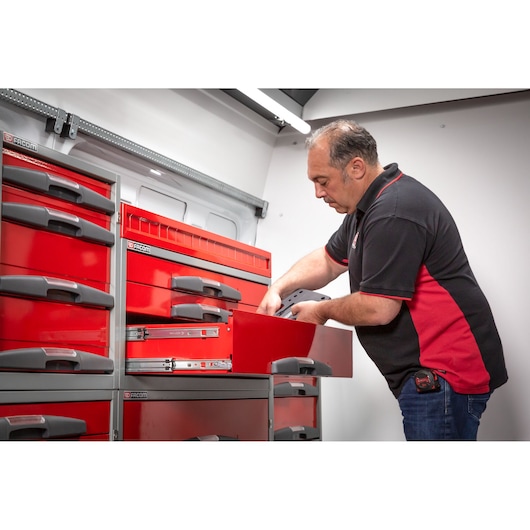 A person wearing a black and red shirt is organizing items in a red Facom storage drawer system, which is mounted inside a vehicle or workspace. The person's face is blurred for privacy.