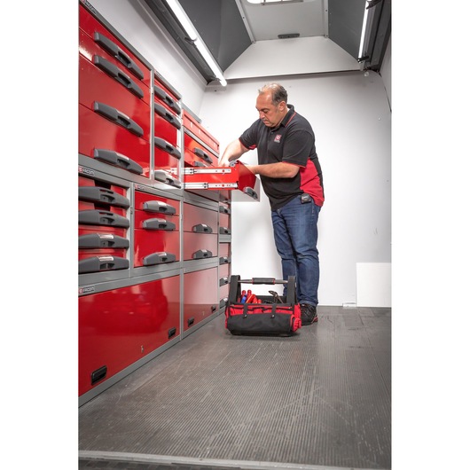 A person is organizing tools inside a red Facom tool storage cabinet in a workshop or vehicle. A black and red tool bag is placed on the floor nearby.