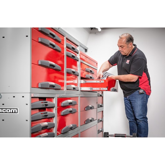 A person standing next to a large red and gray Facom tool storage unit with multiple drawers, opening one of the drawers in a well-lit room.