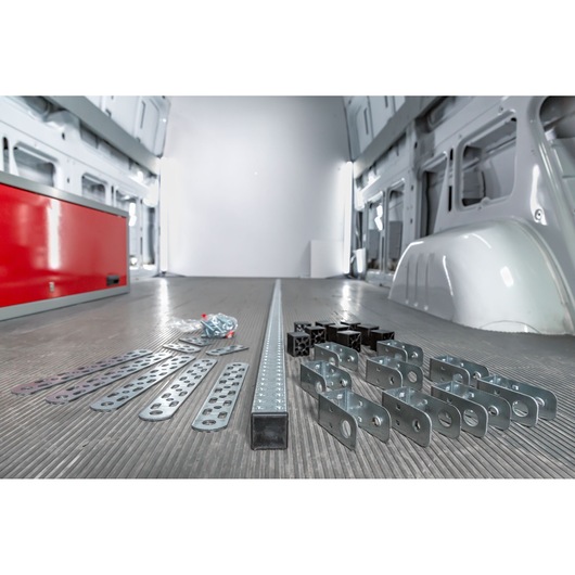 Metal brackets, perforated strips, screws, and plastic connector parts arranged on the floor of a van with grey interior and a red toolbox, photographed for product display.