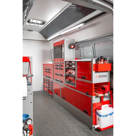 Interior of a service van equipped with Facom red tool storage units and drawers, organized along the side wall. The setup includes multiple drawers, shelves, and compartments, with tools and accessories arranged neatly, and branding visible on the storage. A white water container and various maintenance products are also present.