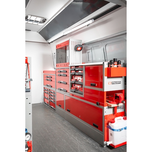 Interior view of a Facom mobile workshop setup inside a van, featuring red and gray tool cabinets, drawers, and shelving units neatly arranged along the side wall. Various tools and accessories are stored, and a white water container is visible at the bottom right.