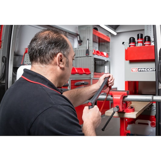 A person using a hacksaw to cut a metal pipe held in a red bench vise, with Facom branded equipment and organized tools in a workshop setting.