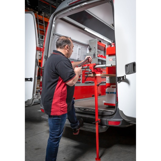 A person is working on a vise attached to a workbench in the back of a van equipped with organized Facom tools and storage modules. The van doors are open and the workspace is designed for mobile tool use.
