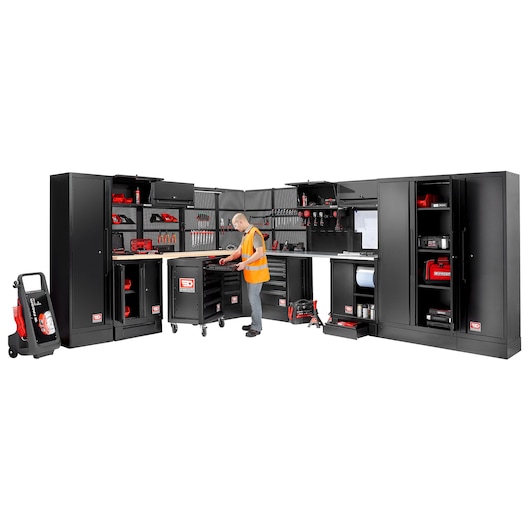A workspace setup featuring multiple black Facom storage cabinets and drawers organized in an L-shape, with various tools and equipment neatly stored. A person in a high-visibility vest is working at the central workstation with tools.