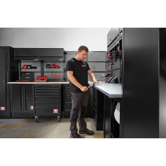 A person is standing in a modern workshop with black Facom storage cabinets, drawers, and workbenches. The individual is working at a metal countertop surrounded by various hand tools and power tools neatly organized on shelves and pegboards.