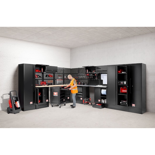A modern workshop setting featuring black modular storage cabinets, tool drawers, and wall-mounted tool racks filled with various hand tools and equipment. A person wearing a safety vest is organizing tools in the central drawer unit. The workspace is clean and well-organized, with a range of red and black tools visible throughout.