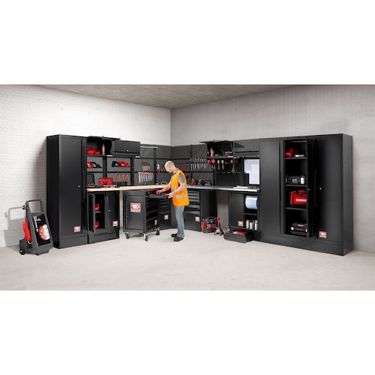 A well-organized workshop workspace featuring Facom black storage cabinets, tool drawers, wall-mounted tool racks, and various red and black tools. A person in an orange safety vest is working in the center, using a rolling tool cabinet. The setting is clean, modern, and industrial.