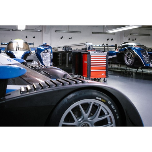 A red Facom JET.7M3A tool cabinet with multiple drawers is positioned in a professional garage, surrounded by blue racing cars on stands and a clean workshop environment.