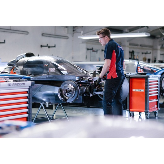 A mechanic works on a racing car in a garage, with two red and black Facom tool cabinets visible nearby. The mechanic is wearing a navy blue and red uniform. The garage environment is well-lit, and the car appears to be under repair or maintenance.