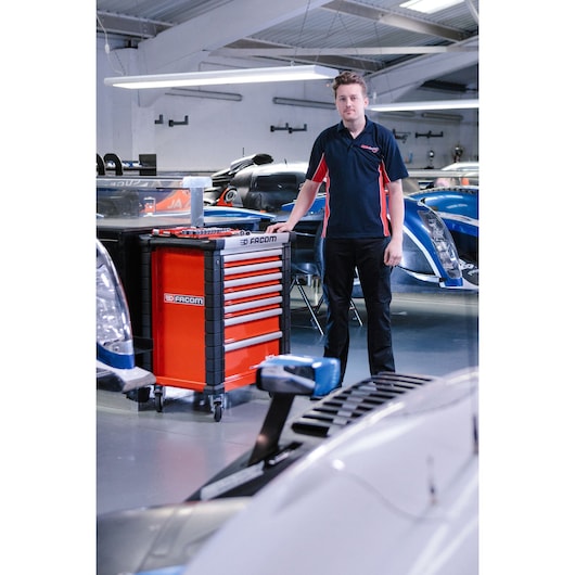 A person standing next to a red Facom tool cabinet with multiple drawers in a garage or workshop environment, surrounded by sports cars. The person's face is blurred.
