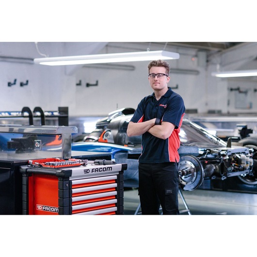 A Facom JET.7M3A tool chest with drawers is shown in the foreground, with various tools displayed on top. A person wearing a navy and red polo shirt stands beside it with arms crossed. In the background, there is a partially assembled race car in a garage or workshop setting.