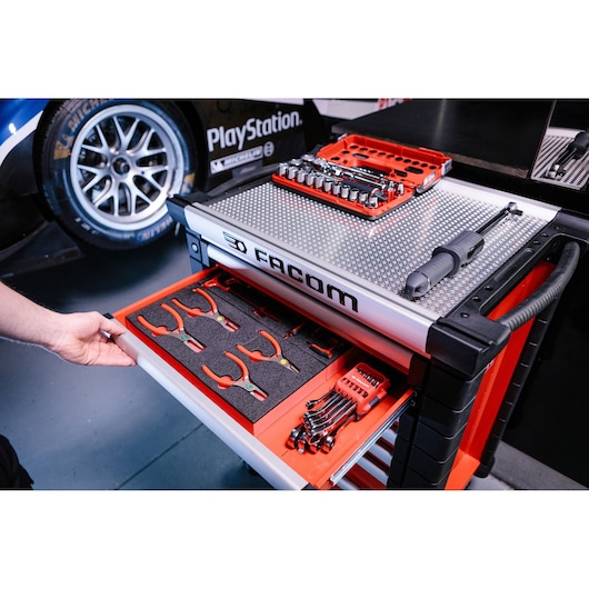 A Facom tool cabinet with various hand tools, including pliers, sockets, wrenches, and a screwdriver, organized in foam trays. The cabinet is placed next to a racing car in a garage setting.