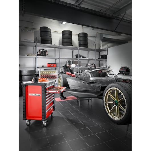 A modern garage or workshop featuring a Facom red tool chest on wheels in the foreground with open drawers displaying organized tools. In the background, a person is working on a race car next to shelves stacked with tires and various equipment.