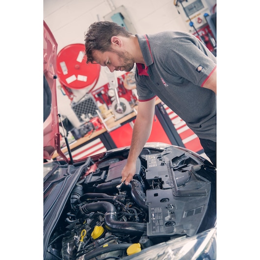 Photo of person inspecting car engine in workshop with Facom R_181 shirt, red tool cabinets in background.