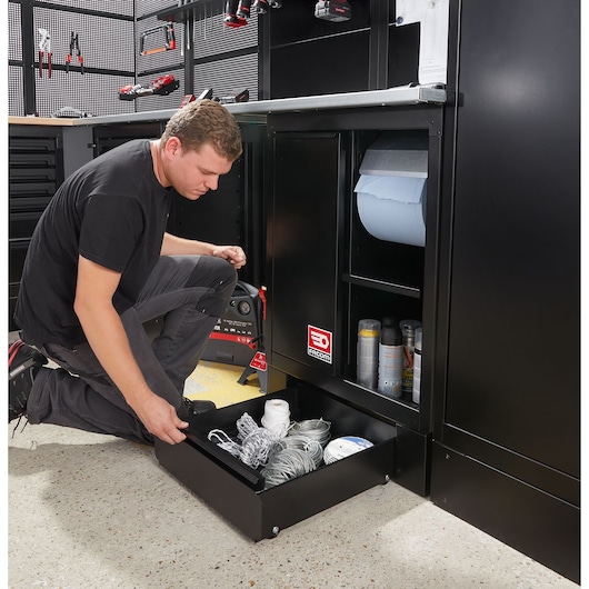 Facom RWS2-DRAWERBS black workshop cabinet drawer open with tools and supplies, person kneeling in front.