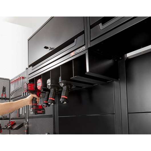 Facom RWS2-POWRACKBS black metal power tool rack mounted under cabinet, four drills stored, photo side angle.