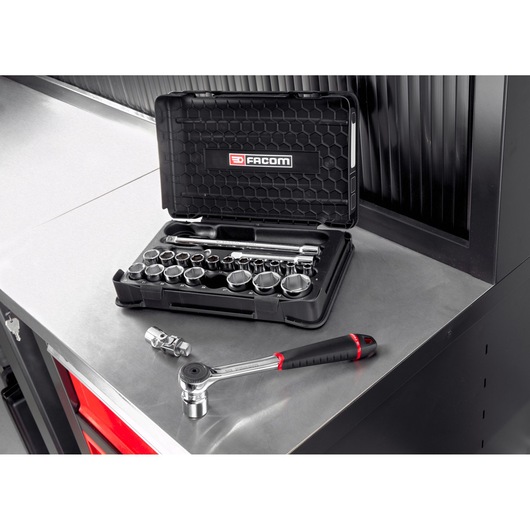 A Facom socket wrench set in a black case displayed on a metal workbench. The case is open, showing various sockets and accessories. A ratchet wrench and socket extension are placed next to the case.