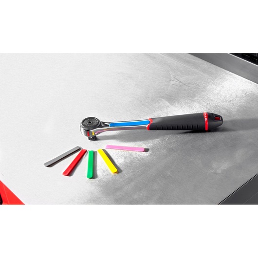 A Facom ratchet wrench with a black and red handle is placed on a metallic surface alongside six colored plastic tags in grey, red, green, yellow, pink, and black.