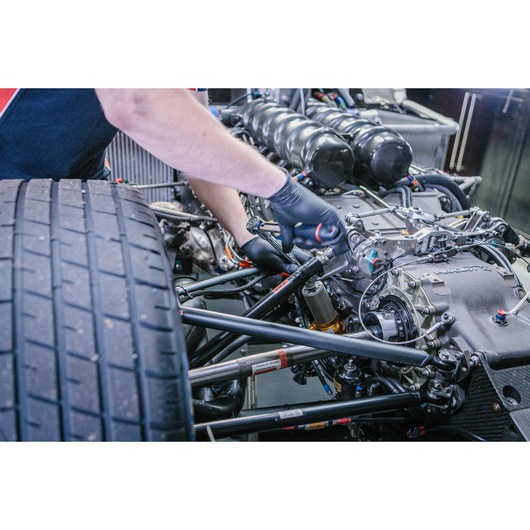 A person wearing black gloves uses a tool to work on the engine and mechanical parts of a racing car.