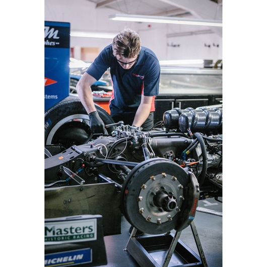 A mechanic working on the exposed engine and braking system of a racing car in a garage. The mechanic is wearing gloves and a dark polo shirt, and is using tools to adjust components of the car.
