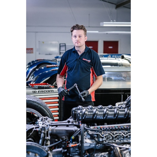 A person wearing a dark blue and red Facom polo shirt and black gloves is standing in a workshop, holding a ratchet wrench. There are car parts and tools visible in the background and foreground.