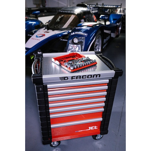 A red and black Facom tool chest with multiple drawers is shown on wheels, with a socket and ratchet set placed on top. The tool chest is situated in a garage with a race car in the background.