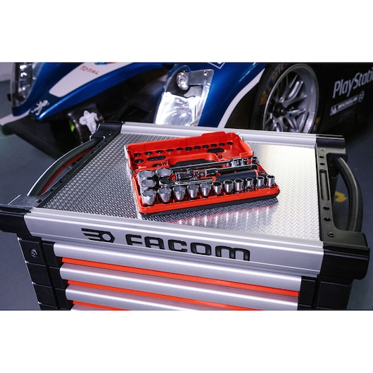 A Facom tool chest with a red socket set and ratchet placed on top, photographed in a workshop with a blue racing car in the background.