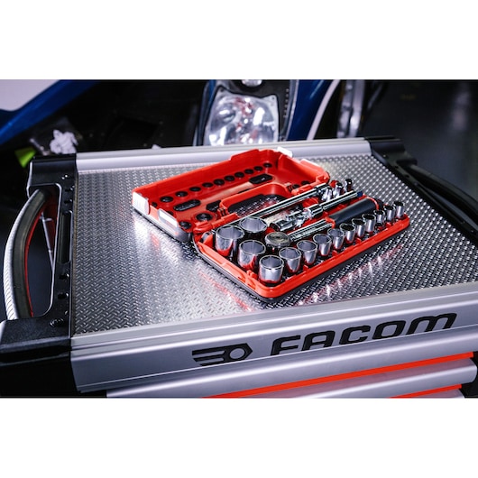 A red Facom socket and ratchet tool set box is placed open on top of a metallic tool chest with a textured surface, displaying various sockets and ratchet tools organized inside.