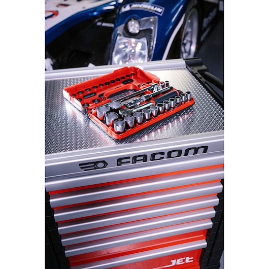 A red Facom socket set with various sockets and tools is displayed on top of a Facom multi-drawer tool chest, with part of a race car visible in the background.