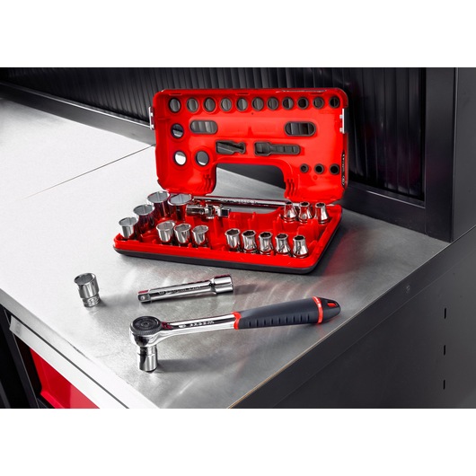 A red Facom socket and ratchet set is displayed open on a metal workbench, showing multiple sockets, extensions, and a ratchet tool. A few pieces are placed nearby on the bench.