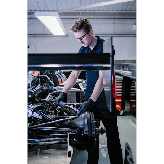A mechanic wearing black gloves uses a wrench to work on the suspension of a race car in a garage. Tool cabinets and various equipment are visible in the background.