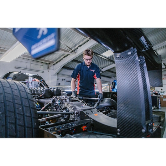 A mechanic in a workshop wearing a navy blue shirt with red accents is working on the exposed engine and rear assembly of a race car. The image shows detailed car components and large rear tires, with the mechanic's face blurred for privacy.