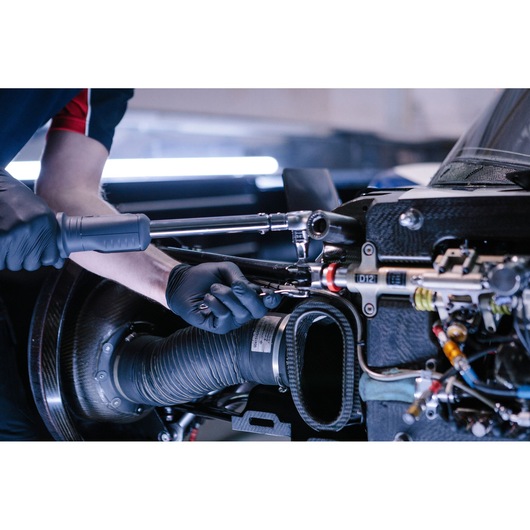 A mechanic wearing black gloves uses a Facom ratchet and socket tool to work on a complex part of a car engine, with visible cables and components.