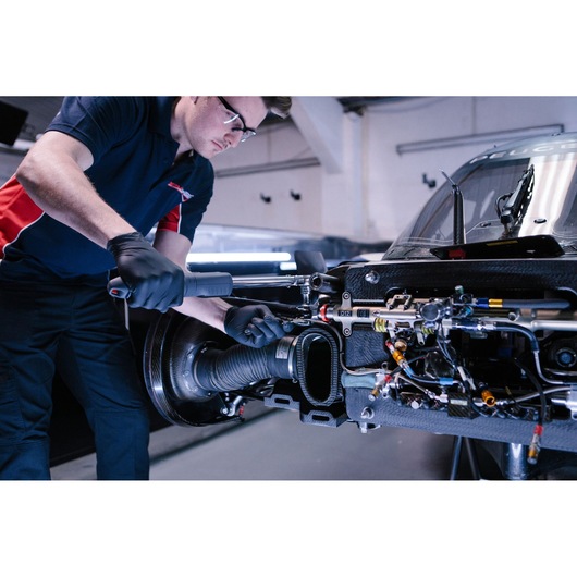 A mechanic wearing black gloves and a blue and red shirt uses a Facom ratchet wrench to work on the exposed engine components of a race car in a garage.
