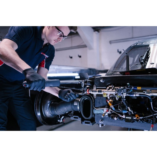 A mechanic wearing black gloves uses a Facom ratchet wrench to work on the exposed inner components of a car in a workshop.