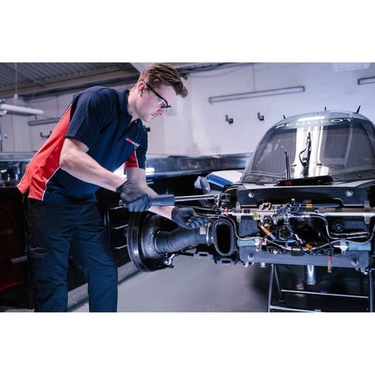 A person wearing a navy and red shirt and black gloves is working on the exposed engine area of a race car inside a garage. The person's face is blurred.