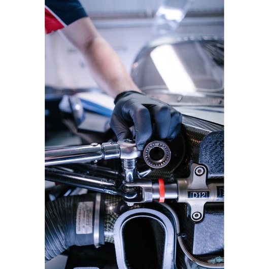 A close-up of a person wearing a black glove and using a Facom ratchet wrench on a mechanical part, likely part of a vehicle engine or machinery.