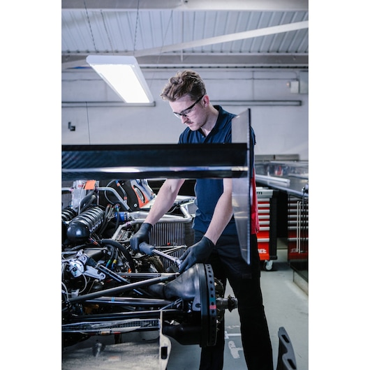 A person with blurred face wearing black gloves uses a tool to work on the engine of a race car inside a workshop, with tool cabinets visible in the background.