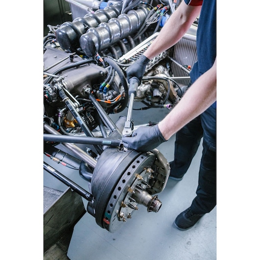 A person wearing black gloves uses a Facom torque wrench to adjust a component on a car's suspension and brake assembly, with an exposed engine visible in the background.