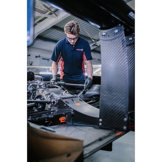 A person wearing a black and red Facom shirt and black gloves is working on the engine or internal components of a vehicle in a workshop. The person's face is intentionally blurred.
