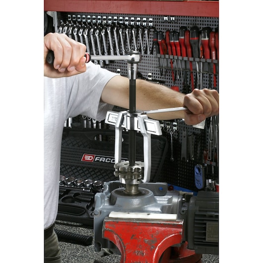 Facom three-jaw gear puller U_232-340 in use on machine part in workshop, mechanic's hands operating tool.