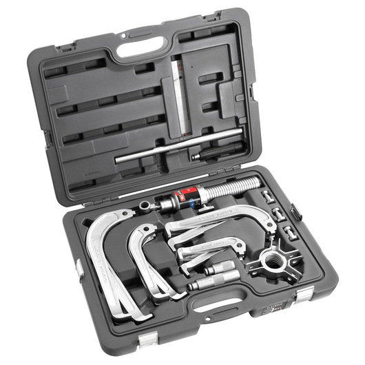 A black plastic case containing a Facom hydraulic puller tool set, including various metal puller arms, a hydraulic cylinder, extension rods, and fittings, organized neatly in fitted compartments.