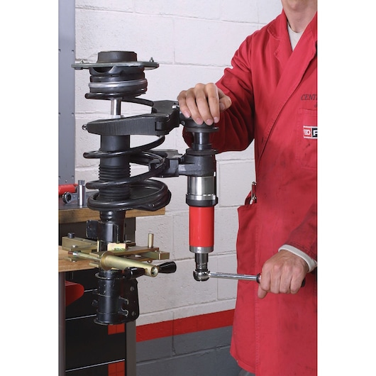 Facom U_89M2 automotive spring compressor in use by technician, workshop setting, side angle.